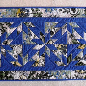 May include: A blue and white quilted table runner with a geometric pattern. The runner has a floral print border.