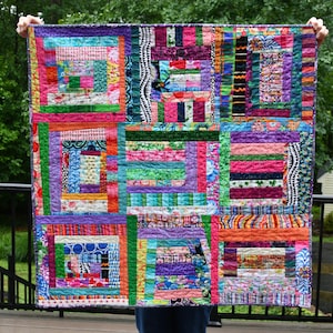 May include: A colorful patchwork quilt with a vibrant mix of fabrics in various patterns and colors, including pink, green, purple, and orange. The quilt features a log cabin design with rectangular blocks and a stitched border. The quilt is held up against a green background.