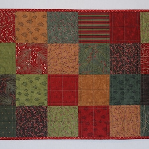 May include: A patchwork table runner with a red, green, and brown color scheme. The runner is made of various fabrics with different patterns, including pine trees, berries, and branches.
