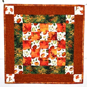 May include: A brown and white quilted table runner with a patchwork design featuring fall leaves and pumpkins. The runner has a scalloped edge and is perfect for a Thanksgiving or autumn-themed table.