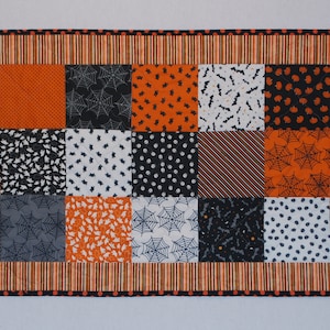 May include: A rectangular Halloween-themed table runner. The center features a patchwork design with orange, black, and gray squares, each with a different pattern, including pumpkins, bats, and spiderwebs. The border is striped with orange and black.