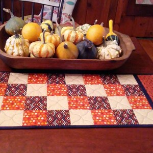 May include: A quilted table runner with a checkered pattern in orange, white, and black. The runner is on a wooden table with a bowl of gourds and a small pumpkin figurine.
