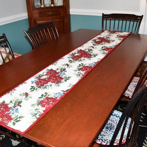 May include: A rectangular wooden dining table with a floral table runner. The runner features a white background with red flowers, green leaves, and blue accents. Dark wooden chairs surround the table, and a rug is visible beneath.