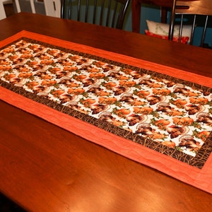 May include: A Thanksgiving table runner with a pattern of turkeys and pumpkins on a white background. The runner has a brown and orange border.