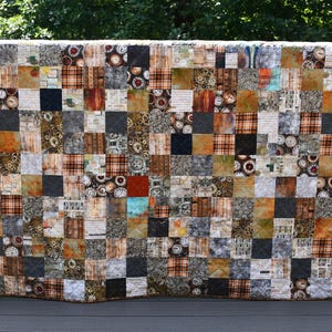 May include: A rectangular patchwork quilt composed of numerous small square fabric pieces in shades of brown, black, gray, and orange. The quilt features a variety of patterns including plaid, clock faces, gears, and script text on a dark background.