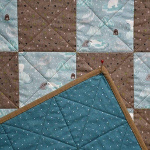 May include: A blue and brown patchwork quilt with a white pin in the corner. The quilt is made of squares with different patterns, including polar bears and icebergs.