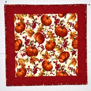 May include: A red quilted table runner with a white background featuring a repeating pattern of orange pumpkins, red berries, and brown leaves.