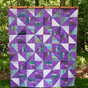 May include: A handmade quilt with a geometric pattern of purple, white, and teal triangles. The quilt is displayed outdoors, hanging from a wooden rod. The design creates a visually appealing patchwork effect, perfect for a bedroom or living room.