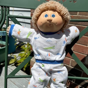 May include: A Cabbage Patch Kid doll wearing a white and blue pajama set with a green alligator print. The doll has blonde yarn hair and blue eyes. The doll is sitting on a green metal chair.