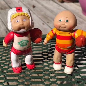 May include: Two Cabbage Patch Kids figurines dressed in sports uniforms. One is wearing a red and white football uniform with a helmet and holding a football. The other is wearing a yellow and red striped shirt and red shorts and holding a football.