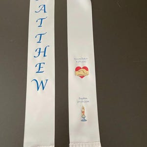 May include: White satin ribbon with blue embroidered lettering spelling "MATTHEW". The ribbon has a white fringe on the bottom. The right side of the ribbon has a red heart with two hands inside, and the text "Congratulations 8-01-2023". Below the heart is a candle with the text "Baptism 29-01-2014".