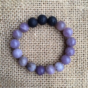 May include: A circular beaded bracelet featuring a series of smooth, round, light purple and lavender-colored stones interspersed with three textured, porous, matte black volcanic lava beads positioned at the top of the elasticized jewelry piece.