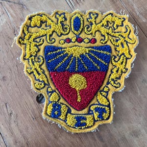 May include: A vintage, embroidered patch in a shield shape. The patch features a yellow border with ornate designs, surrounding a central emblem with red, blue, and yellow elements. A banner at the bottom has blue text on a yellow background.