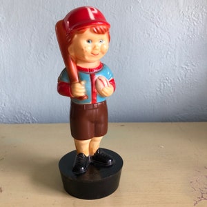 May include: A plastic toy baseball player figurine with a red hat, blue shirt, brown shorts, and a baseball and bat in hand. The figurine is standing on a black base.