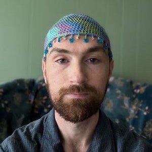 May include: A man wearing a blue, green, and purple crocheted hat with blue beads around the brim. He is wearing a blue button-down shirt.