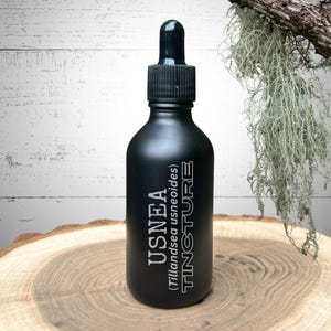 May include: A black tincture bottle with a dropper, labeled "USNEA Tincture". The bottle sits on a wooden surface, with a backdrop of white wood and hanging greenery. The text on the bottle is in white.
