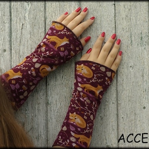 May include: A pair of burgundy arm warmers with a repeating pattern of orange foxes, leaves, and berries. The cuffs are a darker burgundy. The word "ACCESSORI" is printed in white at the bottom right.