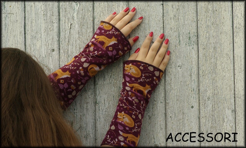 May include: A pair of burgundy arm warmers with a repeating pattern of orange foxes, leaves, and berries. The cuffs are a darker burgundy. The word "ACCESSORI" is printed in white at the bottom right.