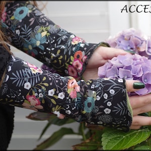 Könnte beinhalten: Ein Paar schwarze Armwärmer mit Blumenmuster und grünem Saum. Die Armwärmer sind aus einem weichen, dehnbaren Stoff gefertigt und zeigen eine Vielzahl von bunten Blumen und Blättern. Die Armwärmer werden von den Händen einer Person gehalten, die auch dunkelgrünen Nagellack trägt.