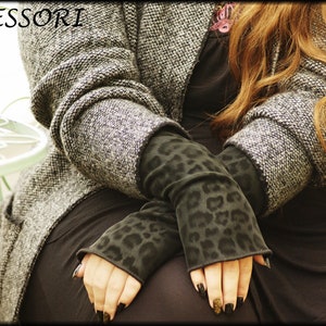 Könnte beinhalten: Ein Paar schwarze Fingerlose Handschuhe mit Leopardenmuster. Die Handschuhe werden von einer Person getragen, die einen grauen Pullover und eine schwarze Hose trägt.