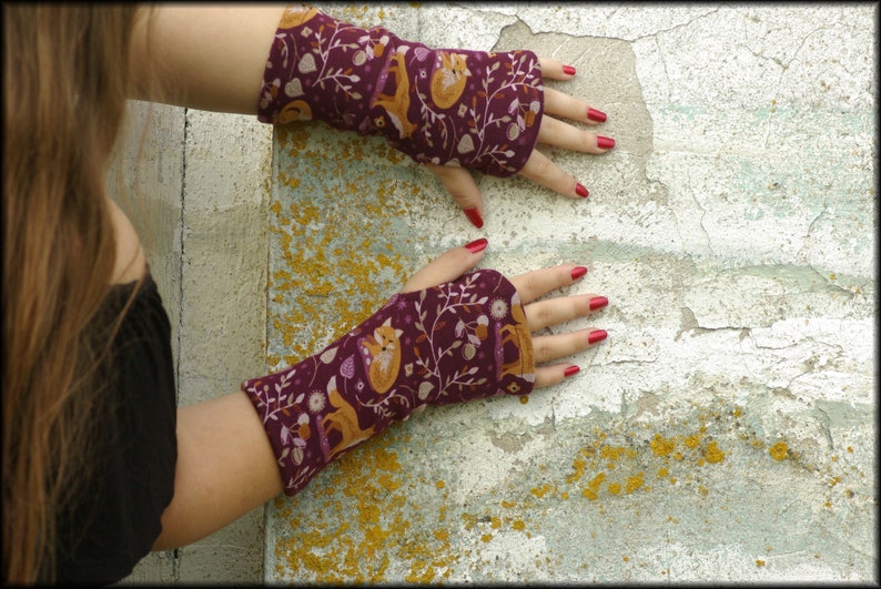 May include: Fingerless gloves with a burgundy background and a repeating pattern of foxes, leaves, and acorns in shades of orange and brown. The gloves are made of a soft fabric and are designed to cover the hand and wrist, leaving the fingers exposed.