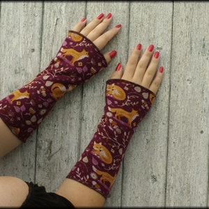 May include: A pair of burgundy arm warmers with a repeating pattern of orange foxes, white flowers, and leaves. The arm warmers have a brown trim and are displayed on a light wood surface.
