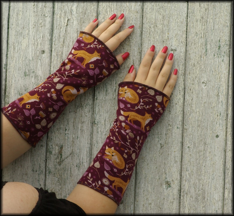 May include: A pair of burgundy arm warmers with a repeating pattern of orange foxes, white flowers, and leaves. The arm warmers have a brown trim and are displayed on a light wood surface.