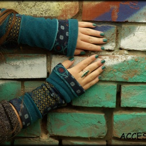 Könnte beinhalten: Ein Paar Armwärmer mit tealblauem und braunem Muster und einem Ring am Finger der Hand, die die Armwärmer trägt. Die Armwärmer befinden sich an einer Backsteinwand mit grüner und weißer Farbe.