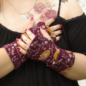 May include: Fingerless gloves with a burgundy background and a repeating pattern of foxes, leaves, and flowers in gold, white, and brown. The gloves are made of a soft fabric and are worn on the hands.