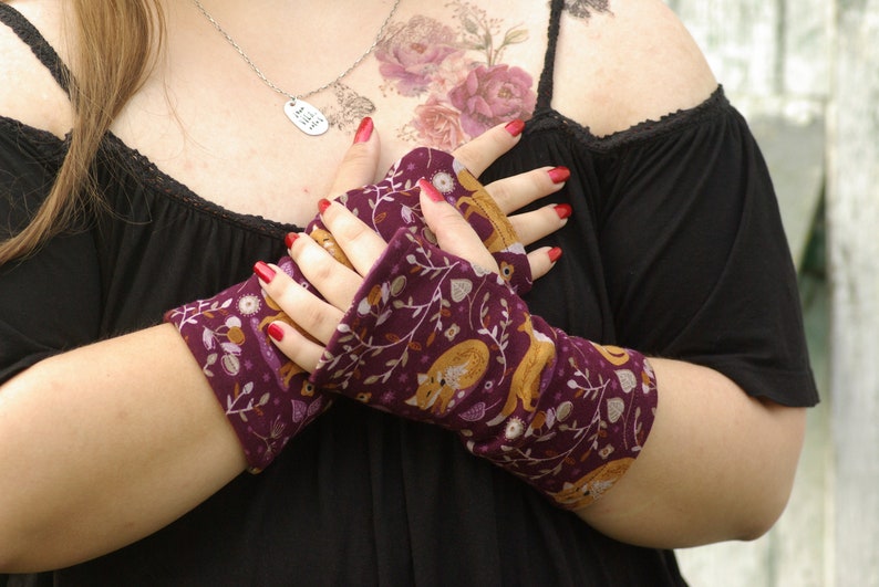 May include: Fingerless gloves with a burgundy background and a repeating pattern of foxes, leaves, and flowers in gold, white, and brown. The gloves are made of a soft fabric and are worn on the hands.