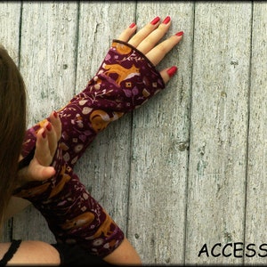 May include: Burgundy arm warmers with a repeating pattern of foxes, leaves, and flowers. The arm warmers have a dark brown trim and are displayed against a weathered, light-colored wooden background. The word "ACCESSORI" is visible.