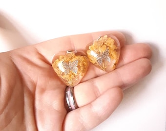 Puede incluir: Dos colgantes de resina en forma de corazón transparente con un dije de mariposa plateada en el interior. Cada colgante está lleno de flores amarillas secas.