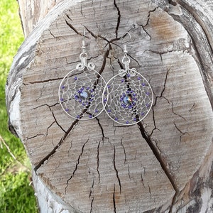 May include: A pair of silver wire earrings with a circular design. The earrings are decorated with small purple beads and a larger iridescent bead in the center of each circle.