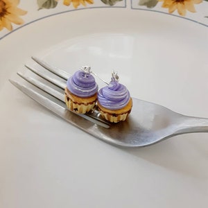 Könnte beinhalten: Ein Paar Ohrringe in Form von Cupcakes in Lila und Gelb. Die Cupcakes haben einen Strudel aus lila Frosting und liegen auf einer silbernen Gabel.