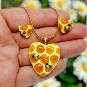 Peut inclure: Un collier en chaîne dorée avec un pendentif en forme de cœur. Le pendentif est en résine jaune et orange et présente un motif en nid d'abeille avec trois petites abeilles. Le collier comprend également une paire de boucles d'oreilles assorties en forme de cœur.