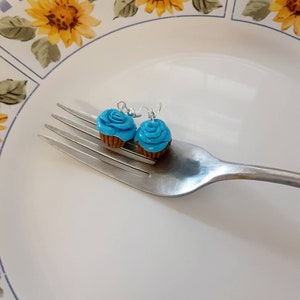 Könnte beinhalten: Ein Paar blaue Cupcake-Ohrringe mit braunen Cupcake-Böden. Die Ohrringe befinden sich auf einer silbernen Gabel, die auf einem weißen Teller mit Blumenmuster liegt.