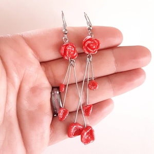 Puede incluir: Un par de pendientes colgantes con abalorios en forma de rosa roja y cuentas rojas más pequeñas que cuelgan de alambres plateados.