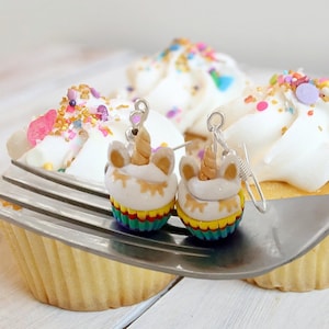 May include: A pair of unicorn cupcake dangle earrings with rainbow frosting and sprinkles. The earrings are silver and have a hook closure.