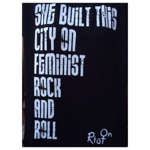 「She Built This City On Feminist Rock And Roll」T シャツは、黒いシャツに白いインクでスクリーン印刷されています。