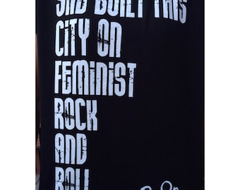 Maglietta She Built This City On Feminist Rock And Roll stampata su maglietta nera con inchiostro bianco.