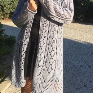 Puede incluir: Un cárdigan gris claro, de manga larga y abierto por delante. El cárdigan presenta un patrón de punto calado detallado con un dobladillo festoneado. Las mangas son largas y el cárdigan llega hasta la mitad de la pantorrilla. La prenda está hecha de un material suave, posiblemente lana.