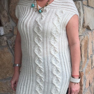 以下が含まれることがあります： クリーム色のノースリーブニットチュニック。前面にケーブルニットのデザイン。チュニックはリブ編みのテクスチャとラウンドネックライン。黒のレギンスの上に着用。ネックレスとブレスレットも見える。