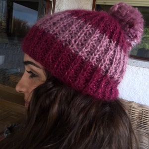 Puede incluir: Un gorro de invierno tejido en tonos rosa y burdeos, rematado con un pompón a juego. El gorro tiene un borde acanalado y un patrón de rayas, con un brillo sutil en todo el hilo. El gorro está diseñado para el clima frío.