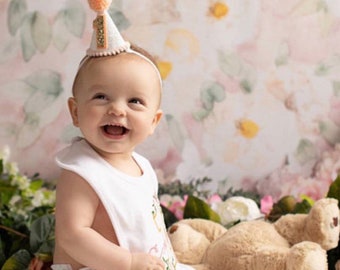 May include: A baby girl wearing a white onesie with a pink and gold embroidered design and a white party hat with a pink pom-pom on top. The baby is sitting on a white surface with a teddy bear beside her. The background is a floral pattern.