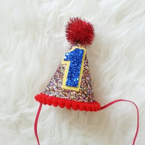 May include: A gold glitter party hat with a red pom-pom on top. The hat has a blue felt number "1" on the front with a yellow outline.