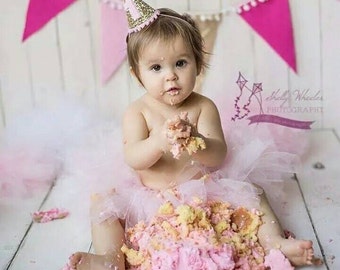 May include: A baby girl wearing a pink tutu and a gold glitter birthday hat is sitting in front of a pink and white cake. She is covered in frosting and has a look of pure joy on her face.