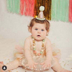 May include: A baby girl wearing a green and gold glitter party hat with the number "1" on it. She is wearing a pink and gold tutu and a multi-colored beaded necklace. She is sitting on a white cake stand with her hands covered in cake frosting.