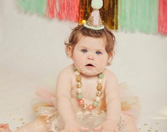 May include: A baby girl wearing a green and gold glitter party hat with the number "1" on it. She is wearing a pink and gold tutu and a multi-colored beaded necklace. She is sitting on a white surface with gold stars scattered around her.