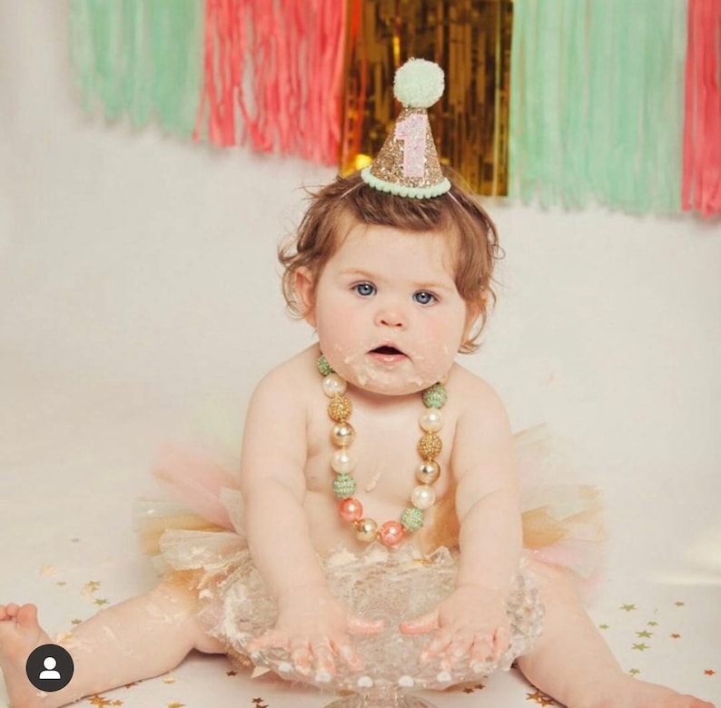 May include: A baby girl wearing a green and gold glitter party hat with the number "1" on it. She is wearing a pink and gold tutu and a multi-colored beaded necklace. She is sitting on a white cake stand with her hands covered in cake frosting.