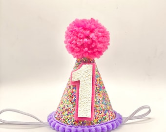 Puede incluir: Un gorro de fiesta de cumpleaños festivo con un pompón rosa. El gorro en forma de cono está cubierto de chispas de colores y tiene un gran número 1 en el centro. Un ribete de pompón morado y una banda elástica completan el diseño.